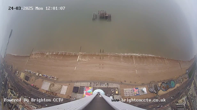 A high-angle view overlooking a beach. The sandy shore is lined with several beach huts and colorful umbrellas. Waves gently lap at the shore, and a worn pier sits partially submerged in the water. The coastline features various attractions, with some people walking along the beach. The scene is captured on a cloudy day, with a timestamp indicating it's noon.
