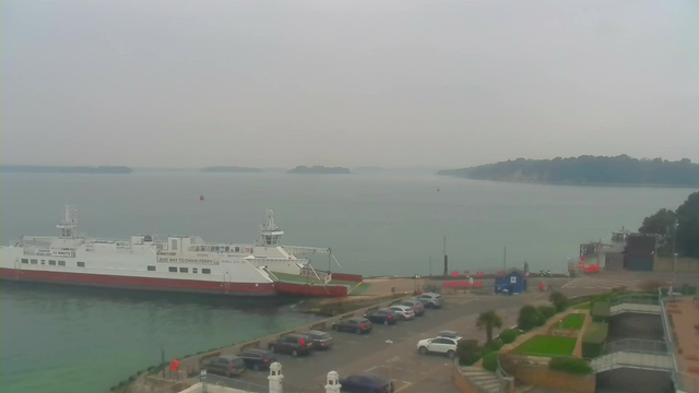 A view of a coastal area featuring a large ferry docked at a terminal. The ferry is white with red accents, and it is located near a parking lot with several cars parked. In the background, there are distant islands and a cloudy sky, creating a hazy atmosphere. There are some trees and green areas along the shore, and a few buildings are visible on the right side of the image. A small boat can be seen on the water.