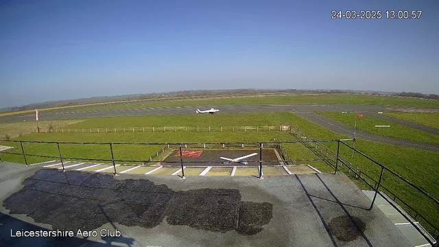 A clear blue sky with no clouds is visible above a wide, green landscape. In the foreground, there is a flat rooftop with a railing, and dark spots indicating wet areas. Below, a small airplane with a red tail is taxiing on a defined runway, surrounded by grass and a wooden fence. The runway has clear markings and stretches into the distance, where it meets more green fields.