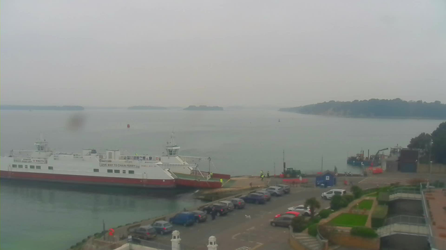 A view of a cloudy harbor scene featuring a large ferry docked at the pier. The ferry is predominantly white with red accents and has the words "Give Way to Chain Ferry" written on the side. In the background, there are several islands and a calm body of water. There are small boats and vessels near the harbor, and a few people in reflective vests can be seen on the pier. A line of parked cars is visible along the waterfront, with a landscaped area containing plants and pathways in the foreground. The overall atmosphere is quiet and overcast.