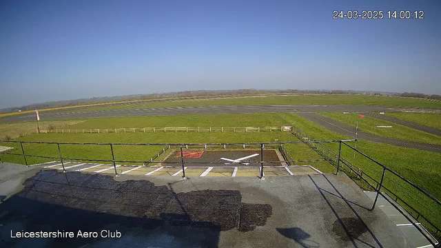 A clear blue sky stretches across the top of the image, with no clouds visible. Below, an airfield is depicted with green grass and a runway. The runway is asphalt and is marked with white lines. In the foreground, there is a rooftop railing and a patch of darker surface, suggesting the building's roof. The image captures a serene, unobstructed view of the landscape with a distant horizon.