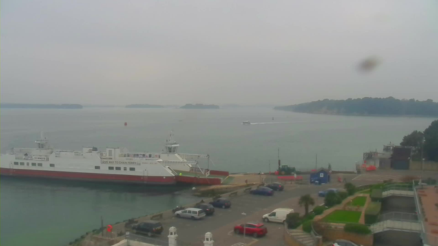 A view of a harbor on a cloudy day. In the foreground, a large white and red ferry is docked at the pier, labeled with safety instructions. Surrounding the ferry, there are several parked cars in a parking area. The water is calm, reflecting the muted colors of the sky. In the distance, several small boats are seen moving across the water, with green scenery and trees visible on the opposite shore. The scene has a gray, overcast atmosphere, suggesting it may be early morning or late afternoon.