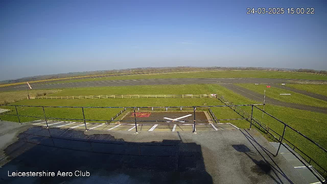 A wide view of an airfield under a clear blue sky. In the foreground, there is a fence and a view from a raised platform or building. Below the platform, a section of the ground is marked with white lines resembling an X, possibly indicating a helicopter landing area. The grassy airfield extends into the distance, with a parking area visible and a small section of an airstrip lined with white markings. The landscape beyond the airfield includes rolling hills and fields. The scene is sunny and bright, showcasing a peaceful aviation environment.