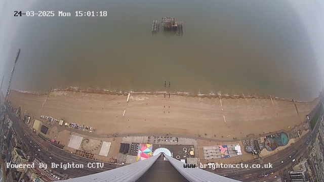 A high-angle view of a beach at Brighton, with the shoreline visible where the sea meets the sandy beach. A pier with a partly collapsed structure is seen in the water, and there are several people walking along the beach. Colorful structures and amenities are situated near the water's edge, along with patches of sand. In the bottom part of the image, there are buildings and a road with cars. The time displayed in the top corner shows Monday, March 24, 2025, at 15:01:18.