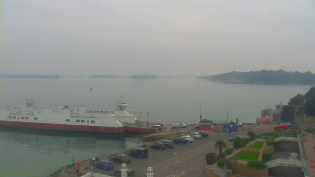 A misty view of a calm bay with a large white and red ferry docked at the pier. In the foreground, there are parked cars along a paved area and some greenery. The water is smooth, reflecting the overcast sky, with a few distant islands visible on the horizon. The scene conveys a quiet, serene atmosphere.