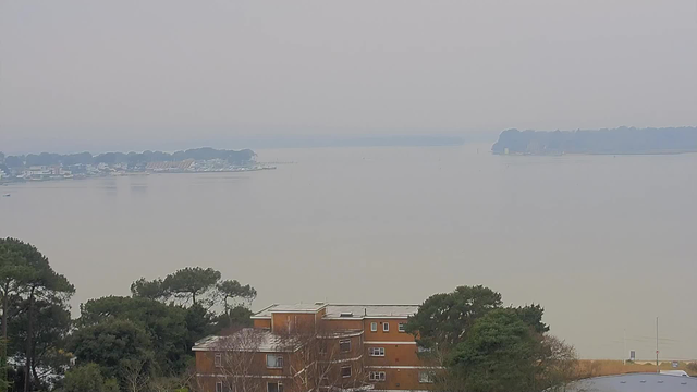 A foggy view of water with a muted grayish-blue sky. In the foreground, there are trees and buildings, including a tan-colored building with several windows. The background features a shoreline with white structures and boats, partially obscured by haze. The water appears calm and reflects the overcast sky.