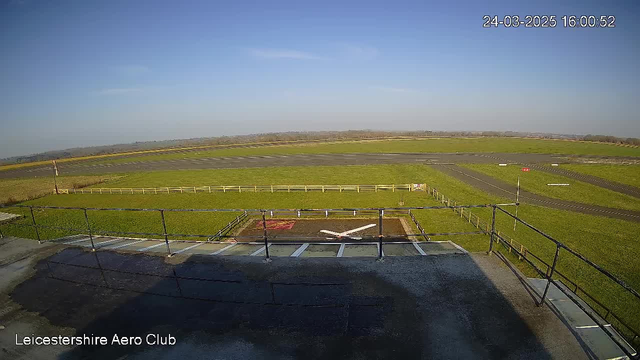 A wide view from a webcam positioned at Leicestershire Aero Club. The foreground features a rooftop railing with a flat surface below, partially wet. In the background, there is a large green field and a runway marked with white lines and a small red flag at the edge. The sky is clear and blue, indicating a bright day.