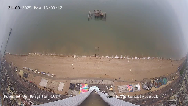 Aerial view of a beach with light brown sand and gentle waves lapping at the shore. The coastline features beachgoers in the distance and a large colorful beach umbrella pattern. There is a pier extending out over the water, and an old, partially submerged structure stands further out at sea. The scene is captured from a high vantage point, giving a broad perspective of the beach and boardwalk area, which includes various structures and vehicles. The sky is overcast, adding a grayish hue to the image.