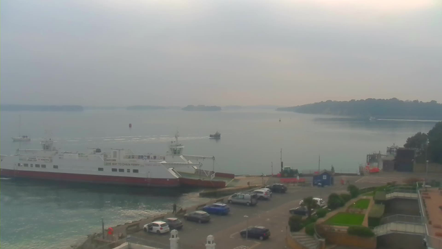 A foggy marine scene showing a large white and red passenger ferry docked at a harbor. In the foreground, a parking area is visible with several cars parked. There are also some people near the water. In the background, a small boat and several distant land masses can be seen on the calm water, which reflects the cloudy sky.
