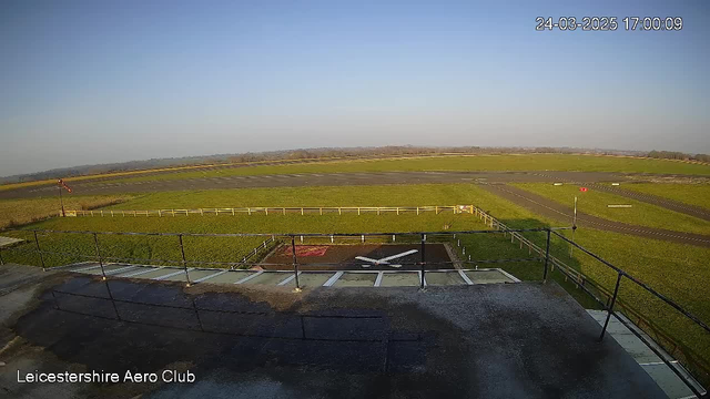 A wide view of an open airfield at the Leicestershire Aero Club during the late afternoon. The sky is clear with minimal clouds. In the foreground, there is a grassy patch with a helicopter landing area marked by a red 'H' symbol and a white diagonal cross. A white airplane silhouette is on the ground near the landing area. To the left, a windsock indicates the direction of the wind, and there are several wooden fences separating different sections of the field. In the background, there are gentle rolling hills visible. The image is timestamped at 17:00 on March 24, 2025.