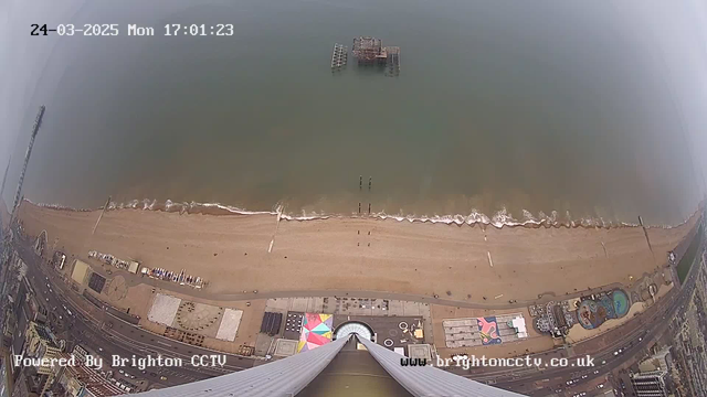 A bird's-eye view of a beach and ocean, taken from a high position. The shoreline shows fine sand and gentle waves lapping at the edge. In the distance, an old wooden pier extends into the water. Below, a colorful amusement area with structures is visible, and there are several small boats along the shore. The image has a timestamp indicating the date and time at the top left corner.