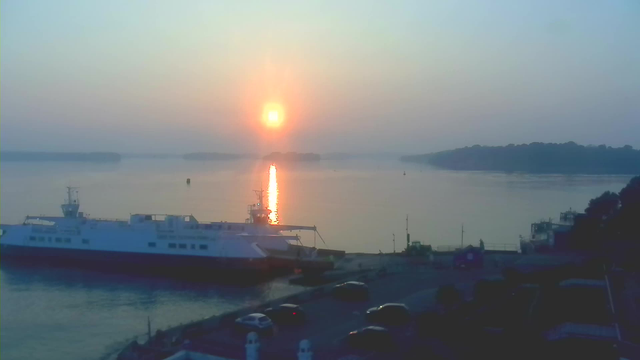 A peaceful scene of a harbor during sunrise. The sun is rising in the sky, casting a warm golden reflection on the calm water. A ferry boat is docked at the pier on the left, with some vehicles parked nearby. The background features distant, hazy landforms, while the sky transitions from blue to soft orange and yellow hues as the day begins.