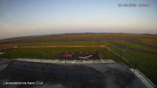 A wide-angle view of the Leicestershire Aero Club's airfield during sunset. The sky is light blue with a hint of orange and pink. In the foreground, there is a safety railing and a tarmac area with a red and white helicopter landing pad, marked with an 'X'. The airfield features a grassy area bordered by a white fence, with a concrete runway leading into the distance. There are few scattered trees and hills visible in the background. The image is timestamped at 18:00:51 on March 24, 2025.