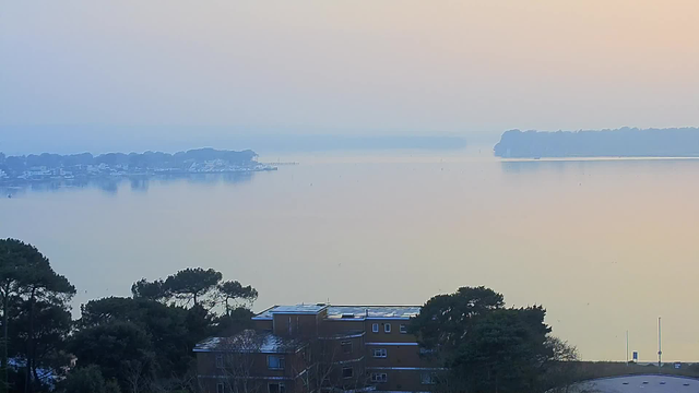 A serene view of a calm lake at sunrise, with a soft gradient of light blue and pale orange in the sky. In the foreground, there are dark green trees and a low building with a flat roof. In the distance, the water reflects the sky's colors, while gentle outlines of land can be seen on either side of the lake. The overall atmosphere is tranquil and foggy, with minimal visibility of distant objects.