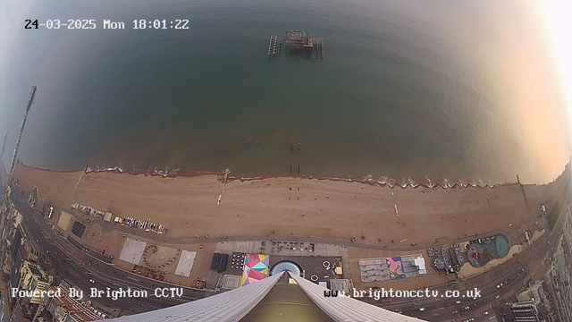 Aerial view of a beach and shoreline during the golden hour. The sand stretches along the coastline, with gentle waves lapping at the shore. A pier extends into the water, and greenish-blue water reflects the sky. There are people walking on the beach and a few scattered structures, including beach chairs and an amusement park area with colorful elements. The bottom of the image shows the edge of a tall building with a large glass area. The date and time are displayed in the top left corner.
