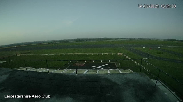 A view from a webcam showing an open field with a runway. The foreground features a flat rooftop area with a helipad marked by a white symbol resembling crossed blades. In the background, there is a large expanse of green grass and a few red and white markers indicating the runway edges. The sky is slightly overcast, with a soft gradient transitioning from light to darker hues. The image is taken from the Leicestershire Aero Club.