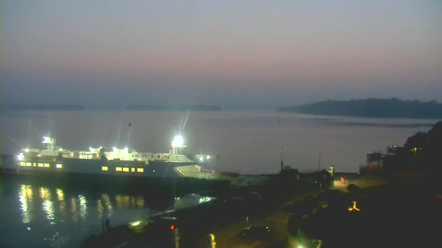 A calm early morning scene featuring a large ferry docked at a port. The ferry is illuminated, with bright lights reflecting on the water. In the background, there are faint outlines of islands and a misty horizon. The sky transitions from light pink to blue as the sun begins to rise. A roadway and some vehicles are visible near the dock, with additional lights punctuating the area. The atmosphere is tranquil, with a sense of dawn approaching.