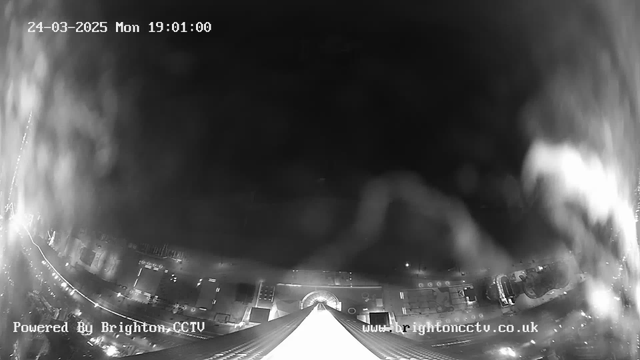 A black and white image from a webcam showing a downward view of a cityscape at night. The top portion of the image displays a pointed roof structure, while the lower portion shows streetlights and building outlines. The scene is somewhat obscured by fog or mist, creating a blurred effect. The time and date are displayed in the top left corner, indicating March 24, 2025, at 7:01 PM. A watermark in the bottom left corner credits "Powered By Brighton CCTV."