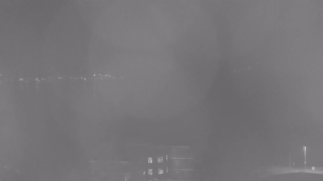 A dimly lit webcam image shows a blurry view of a body of water, with faint lights reflecting on the surface in the distance. In the foreground, partially visible, there are dark shapes that appear to be buildings or structures. The overall atmosphere is dark, with limited visibility.