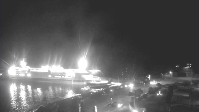 A dimly lit scene shows a large ferry docked at a pier, surrounded by calm water reflecting light. Bright lights on the ferry illuminate the area, casting a glow on the nearby shore. A few vehicles are parked along the road leading up to the dock. The background features faint outlines of structures and trees, with minimal illumination from distant streetlights. The overall atmosphere is quiet and nighttime.