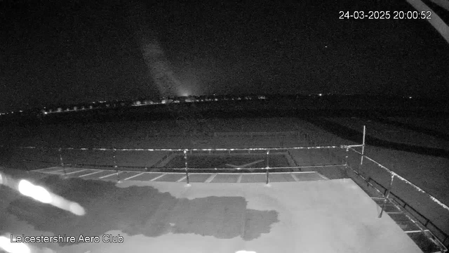 A nighttime view of an empty airfield. The foreground shows a dark surface with a railing along the edge. In the background, there are faint, scattered lights indicating nearby buildings or structures. The sky is mostly dark with a hint of light in the distance, possibly from a rising moon or distant city lights. The timestamp at the top right indicates the date and time as March 24, 2025, 20:00:52.