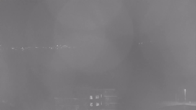 A blurry black and white image showing faint outlines of distant lights and structures. The foreground features a dark building with some illuminated windows, while the background includes indistinct lights that could represent a cityscape or horizon. The overall scene is hazy, suggesting fog or low visibility conditions.