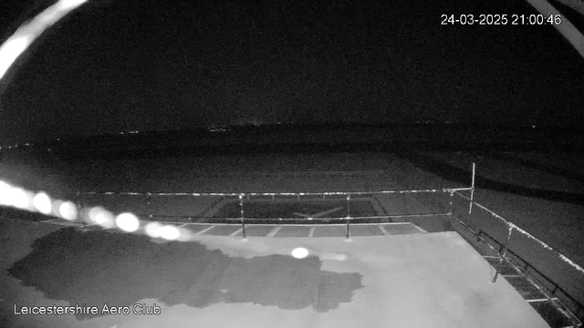 A webcam image shows a dark outdoor area with a slight glow from distant lights on the horizon. In the foreground, there is a flat surface, possibly a runway or platform, partially obscured by shadows. A railing runs along the edge of the platform, indicating a boundary. The scene appears to be set at nighttime. There are no visible objects or people in the immediate area.