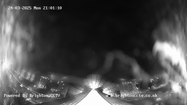 A black and white image from a webcam showing a night scene with low visibility. The view appears to be looking down from a height, possibly a building or structure, with blurred lights visible in the lower part of the image, suggesting a cityscape below. The date and time are displayed in the upper left corner, indicating it is March 24, 2025, at 21:01:10. There is a soft light source in the center, creating a glare that obscures some details in the image. The bottom of the image features text indicating it's powered by Brighton CCTV and a website link.