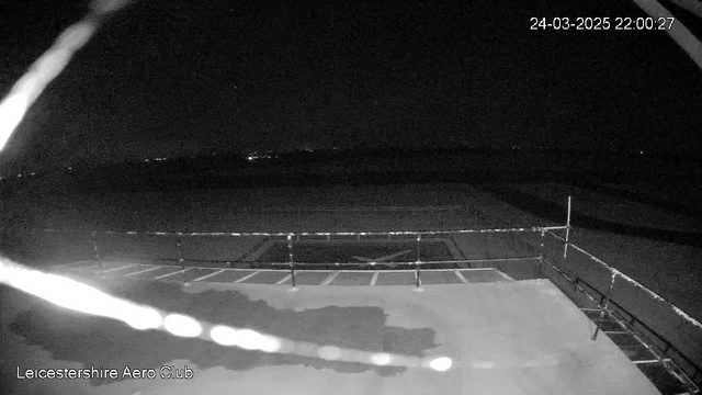 A dark scene showing a flat area, likely an airfield, with a faint outline of a fence or railing in the foreground. There are scattered lights in the distance, indicating potential buildings or vehicles. The image has a timestamp in the upper right corner, indicating the date and time. Overall, the scene is low in visibility with minimal features discernible.