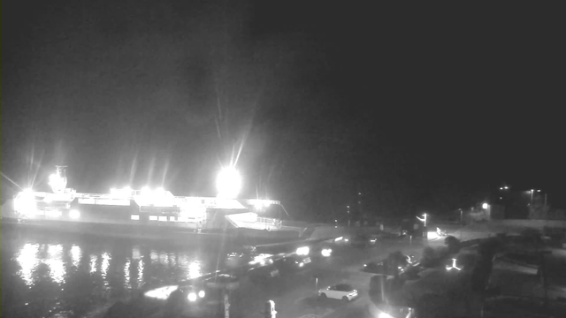 A mostly dark scene shows a large ferry illuminated by bright lights at the dock, reflecting on dark water. In the foreground, several cars are parked along the waterfront road, with some moving towards the right side of the image. There are lights from buildings and street lamps in the background, adding dim illumination to the nighttime setting.