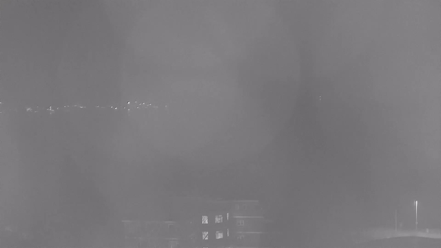 A foggy night scene captured by a webcam. Faint outlines of buildings are visible in the foreground, with a few illuminated windows. In the background, a faint line of lights is discernible, suggesting a distant road or city skyline. The overall image has a grayish tone due to the low visibility caused by fog.