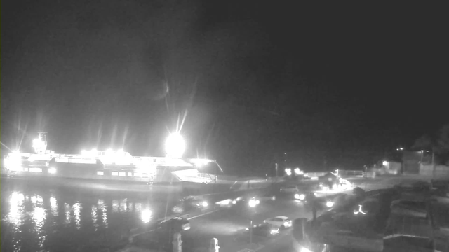 A nighttime view of a harbor area with a large vessel illuminated by bright lights. The reflection of these lights shimmers on the water's surface. In the foreground, a road is visible, with several cars driving past and other lights scattered along the harbor and surrounding buildings. The overall scene is dark but highlighted by the vessel's bright illumination and the headlights of vehicles.