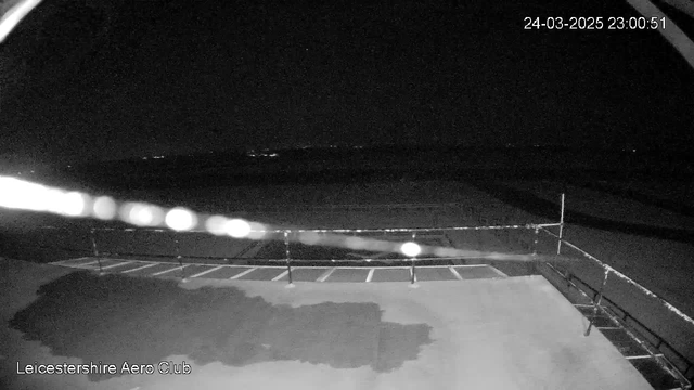 A dark scene captured by a webcam at night. The foreground shows a flat surface with a slight reflection, possibly wet, and a fence in the middle of the frame. There are faint lights in the distant background, suggesting the presence of buildings or vehicles. The timestamp indicates the image was taken on March 24, 2025, at 11:00 PM. The logo for Leicestershire Aero Club is displayed at the bottom left corner.