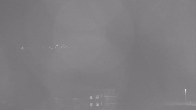 A grayscale image taken from a webcam showing a blurred, indistinct view. In the foreground, there are faint outlines of buildings with some windows appearing slightly illuminated. In the background, a faint line of soft light represents distant lights or structures. The overall clarity is low, suggesting fog or mist affecting visibility.