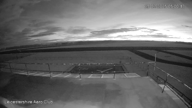 A black and white image from a webcam showing a flat rooftop with a railing on the left side. Below the railing is a large, open space, likely a runway or playing field, with a faint grid pattern visible. The sky above has clouds and is transitioning from dark to light, indicating dawn. The date and time in the corner reads 25th March 2025, 5:00:13. A text label in the bottom left corner states "Leicestershire Aero Club."