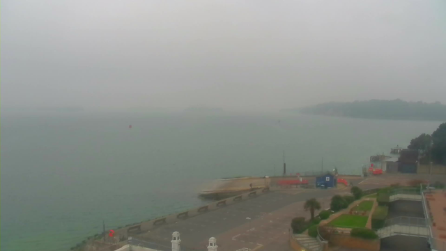 A foggy scene showing a body of water with a faint horizon. In the foreground, there is a light-colored pathway leading to a marina with a few boats, a small blue structure, and red objects near the water's edge. Green grass and plants are present along the pathway. The surroundings appear muted due to the fog, which obscures the view of the water and distant land.