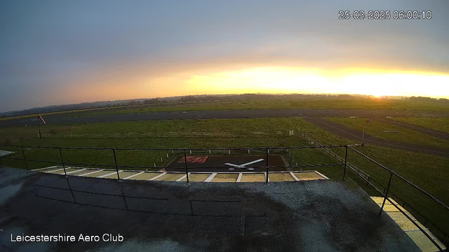 A view from a webcam at the Leicestershire Aero Club, capturing a sunrise over an airstrip. The sky transitions from dark to light, with shades of orange and yellow as the sun rises. In the foreground, there is a flat rooftop with a railing and a small section of the airstrip featuring a white airplane shape marked on the ground. The background shows an open field and a winding path of the runway leading into the distance.