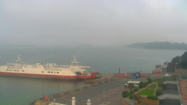 A foggy view of a waterfront scene. In the foreground, a large ferry boat is docked, featuring a white hull with red accents. Nearby, there are several vehicles parked along a waterfront road, with a few decorative plants and grass areas visible. Across the water, a faint outline of land can be seen in the distance, shrouded in mist. The sky is gray, contributing to the overall subdued atmosphere.