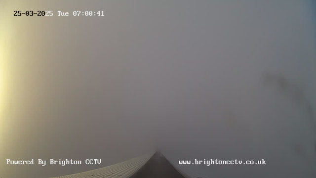 A foggy scene is shown from a webcam, where visibility is extremely low. The image captures the end of a structure leading into the fog, with no distinguishable features visible beyond a few meters. In the corner, there is a timestamp indicating March 25, 2025, at 07:00:41, and a logo that says "Powered By Brighton CCTV" along with the website URL "www.brightoncctv.co.uk." The overall atmosphere is muted with soft colors, primarily gray.