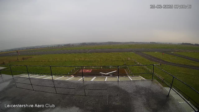 A view of a grassy airfield under a cloudy sky. In the foreground, there is a fenced area with a red and white shaped marking on the ground. A runway is visible in the background, alongside some distant trees and a small hill. The image indicates gloomy weather with overcast skies. The bottom left corner has text indicating "Leicestershire Aero Club" and shows a timestamp of March 25, 2025, at 07:00:46.