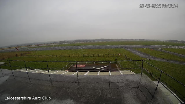 A view from a webcam overlooking a grassy airfield. There are several runways visible, some wet, suggesting recent rain. A small white airplane is situated on a concrete pad in front of a fence. The sky is overcast and gray, with a hint of fog obscuring the horizon. The image is taken from a balcony of the Leicestershire Aero Club, with a railing visible in the foreground. The environment appears quiet and empty, emphasizing the stillness of the scene.