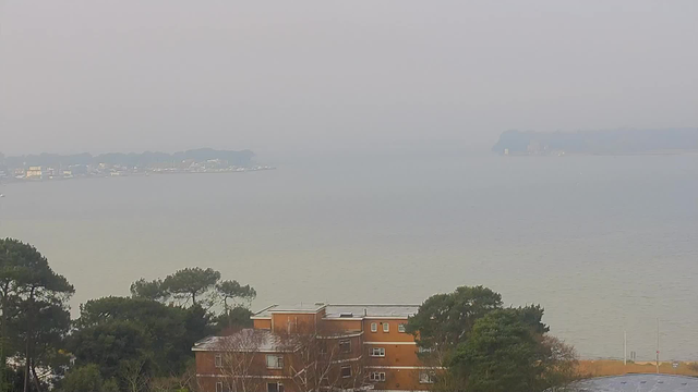 A view of a hazy seascape with water reflecting muted light. In the foreground, there are several buildings, including a brown structure with multiple windows and a flat roof. To the left, greenery suggests trees and shrubs, while the background shows a distant shoreline with a few boats visible. The sky is overcast, contributing to the overall soft and foggy atmosphere.