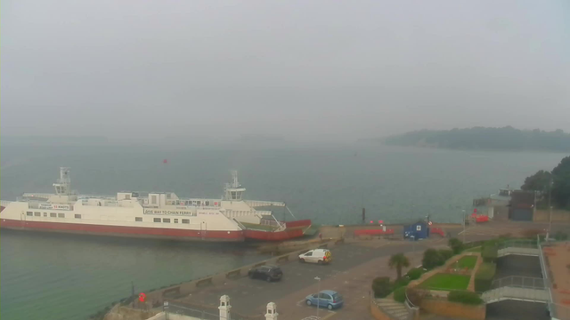 A foggy view of a waterfront scene featuring a large ferry docked at a pier. The ferry has a white body with red accents and is labeled "GIVE WAY TO CHAIN FERRY." In the foreground, there are parked cars on a paved area next to the water. To the right, there’s a path leading to a garden with small hedges and grass. The water is calm, and the background shows land with trees, partially obscured by the fog.