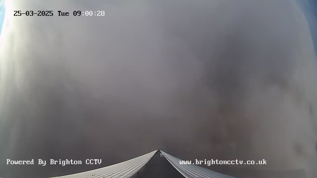A webcam image shows a view from above the roof of a building, looking toward an overcast sky. The scene is mostly covered by thick, gray clouds, obscuring the view of the sky. The building's slanted roof is visible in the lower part of the image, directing upwards towards the clouds. The timestamp at the top indicates the date and time: March 25, 2025, at 09:00:28. The image includes a watermark stating "Powered by Brighton CCTV" at the bottom.