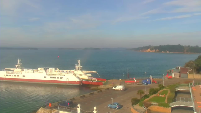 A ferryboat is docked at a harbor, with a calm blue ocean in the background. The ferry is white with a red bottom and has signage visible on its side. Several small vehicles are parked near the harbor area, including a blue car and a white van. There are patches of green grass and a few trees near the dock, and a portion of the shoreline can be seen in the distance, with trees and cliffs. The sky is mostly clear with some light clouds.