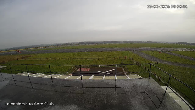 A view from a webcam at Leicestershire Aero Club shows a grassy airfield under a cloudy sky. In the foreground, there is a white airplane silhouette on a red marking, while a flag is visible on a pole nearby. The airfield stretches out in the background, with a worn asphalt runway and a few low fences marking the boundaries. The scene is misty, giving it a subdued atmosphere.