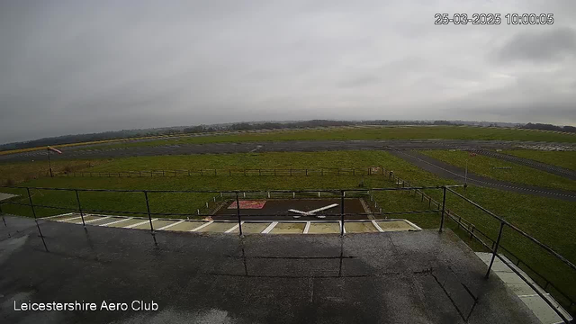 A view from a webcam at the Leicestershire Aero Club overlooking an airfield. The scene is overcast with gray skies and a flat, grassy area. In the foreground, there is a balcony railing and a gravel surface. Below, a large red square is visible along with a white cross marking. The airfield stretches into the distance, showing a solid runway next to grassy patches and a fenced perimeter. There are no visible aircraft in the image. The timestamp in the corner indicates the date and time.