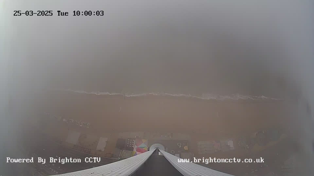 A foggy view from a high vantage point looking down at a sandy beach. The shoreline is faintly visible with gentle waves approaching the shore. Various colorful shapes, possibly beach structures or umbrellas, can be seen in the area near the sand, but visibility is limited due to the fog. The image displays the date and time in the upper left corner, indicating March 25, 2025, at 10:00 AM. The bottom of the image includes a logo and a website link.