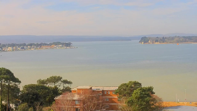 A peaceful landscape featuring a large body of water, with gentle waves reflecting the muted blue sky. On the left side, there is a marina with several boats moored. In the foreground, there are green trees and multi-story buildings with orange and brown exteriors. In the distance, an island with some structures can be seen against a backdrop of rolling hills. The atmosphere is calm, suggesting a clear day with minimal clouds.