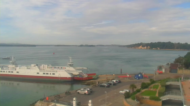 A view of a calm harbor with a large ferry docked at the foreground, painted in white and red. Several cars are parked along the waterfront. In the background, the water stretches out toward distant hills and an island with green trees. The sky is partly cloudy, casting soft light over the scene. To the right, there is a small building, and to the left, a boat is traveling across the water.
